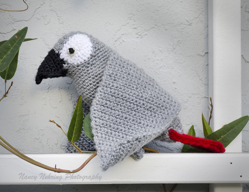 Crocheted African grey parrot amigurumi sitting on an outdoor trellis. Made by Noelle Oguri.